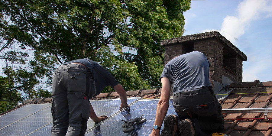 two men wearing blue t-shirt and grey pants installing solar panels on the roof with big tree in the background